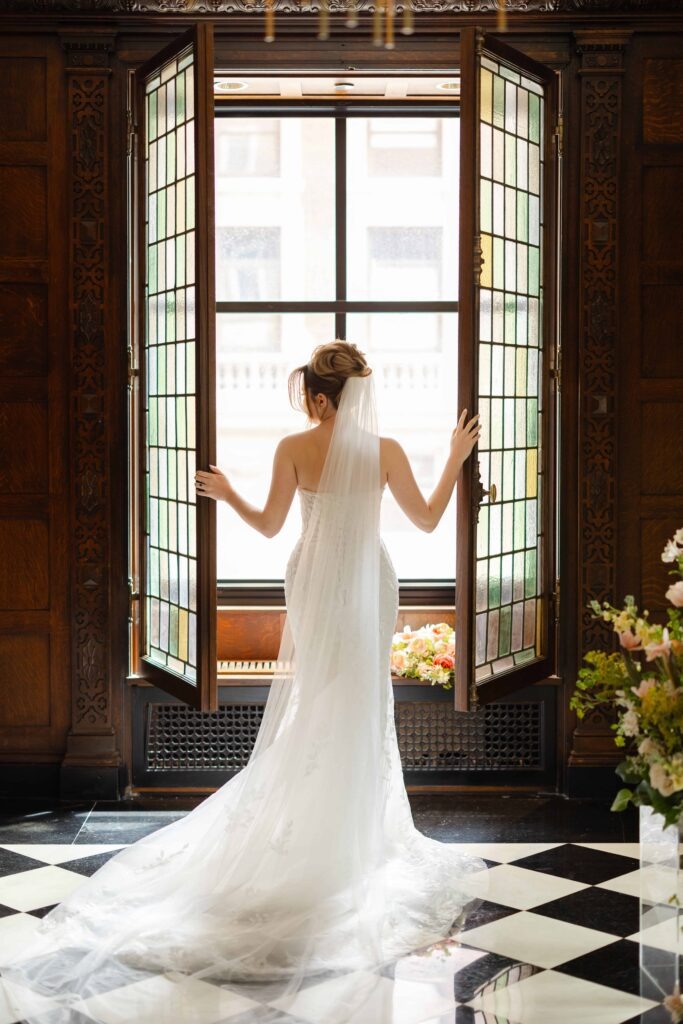 A bride stands facing out of a stained glass window with her veil long behind her at her Blackstone Hotel Chicago wedding
